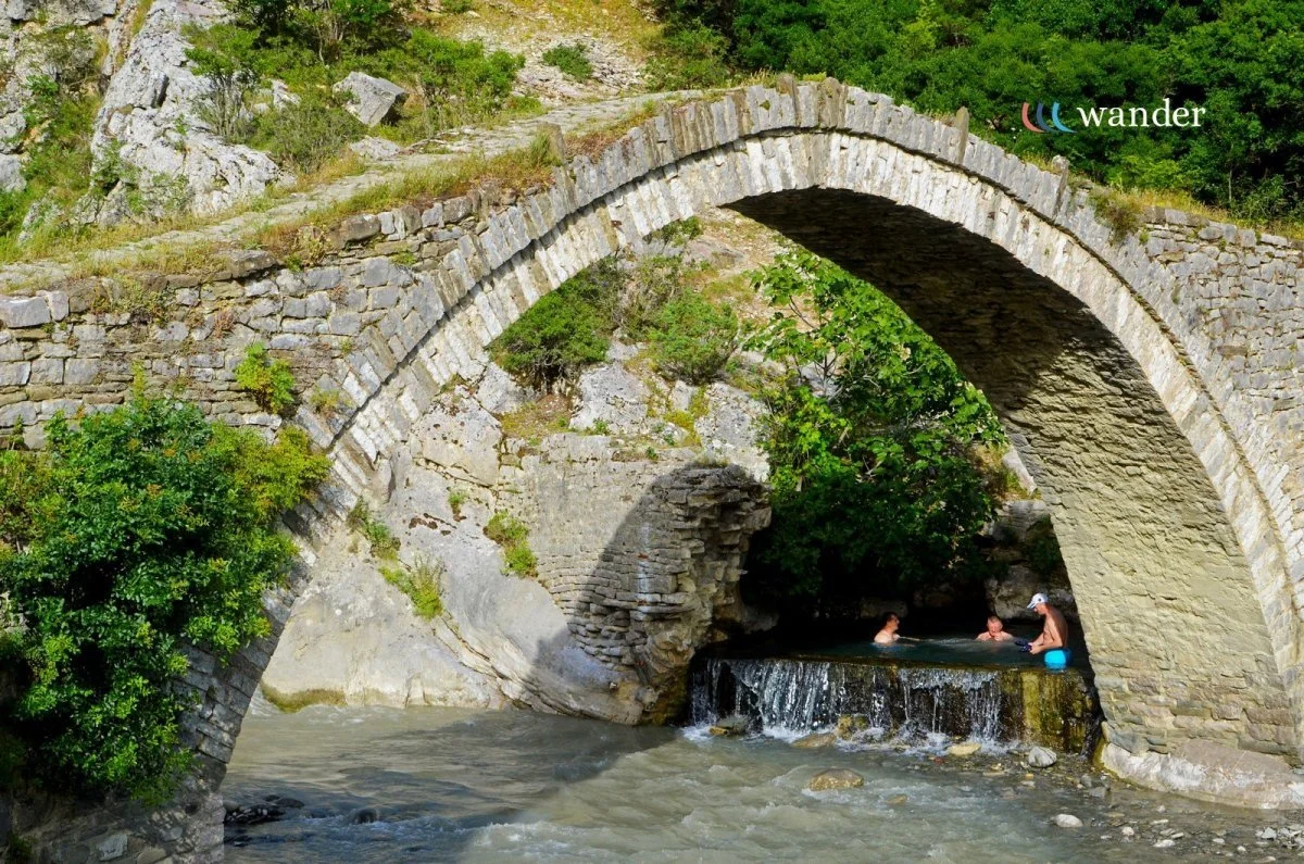 People relaxing in a small hot spring underneath an old stone bridge in a lush green outdoor setting.