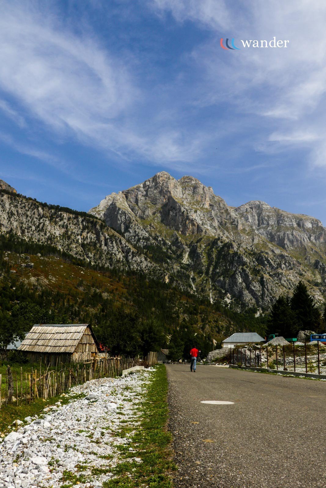 A mountain landscape with a small village, a man in a red shirt walking on a road, a cloudy blue sky, and the logo 'wander' in the top right corner.