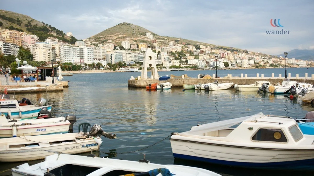 A harbor with several boats docked along the pier, overlooking a city on a hillside with multiple buildings and a mountain in the background, and a logo in the upper right corner reading "wander."