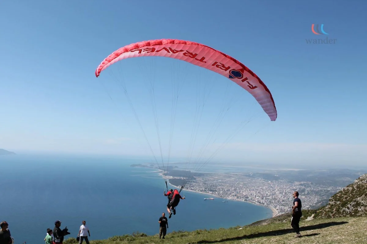 A person with a red jacket and black pants prepares to land from a parachute while others watch on a grassy hillside overlooking a city and ocean.