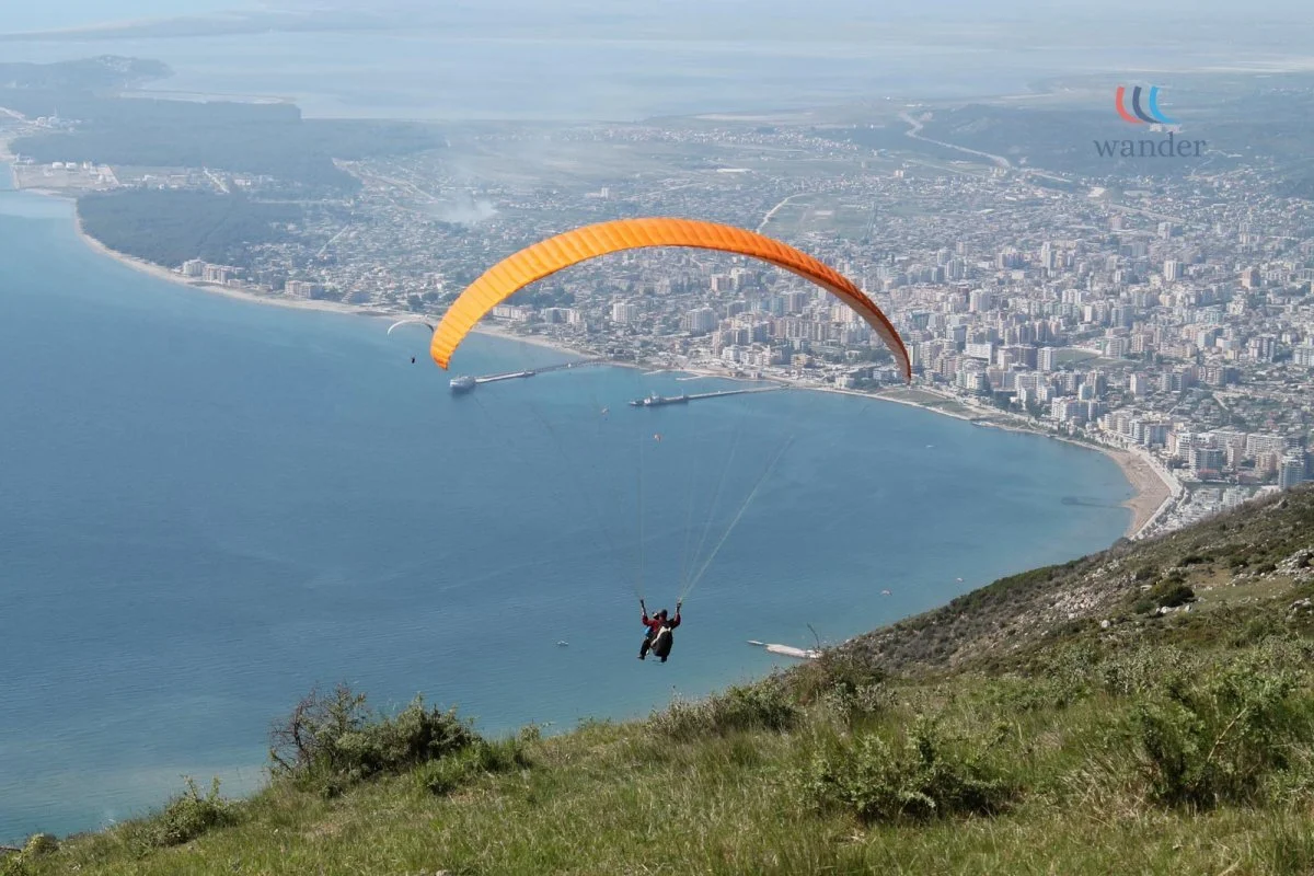 A person paragliding over a coastal city with high-rise buildings along the shoreline, green hills in the foreground, and a large body of water.