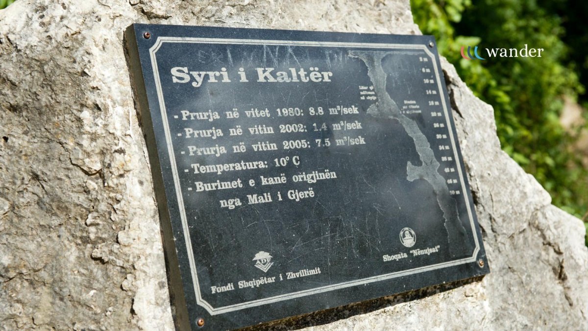Sign mounted on a rock, providing information about the Kalters River, including past flow rates, current temperature of 10°C, and its origin from Mali i Gjerës.