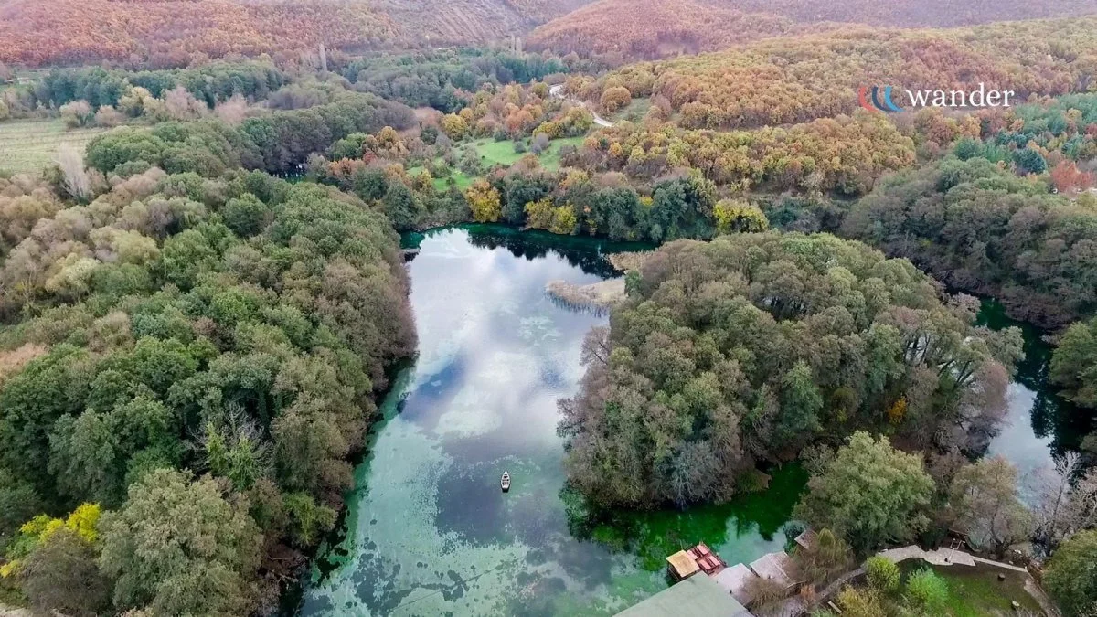 Aerial view of a lake surrounded by dense forest with autumn-colored leaves, a small boat on the water, and a few structures near the shoreline.