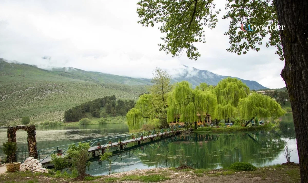 A peaceful lakeside scene with a wooden bridge, lush green trees, and mountains in the background, under a cloudy sky.