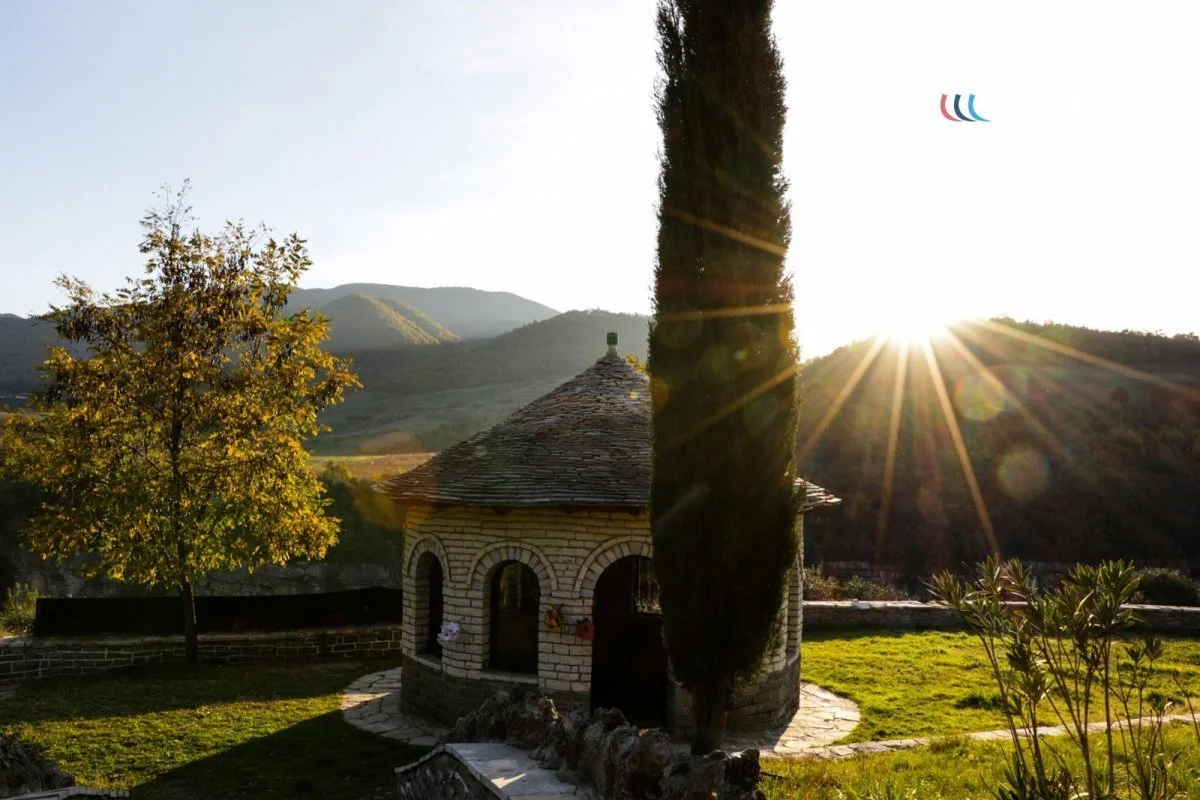 A small stone building with a conical roof, possibly a chapel or a hut, with arched openings. There are trees and bushes nearby, and mountains in the background. The sun is setting behind the mountains, creating a bright glare with sun rays.