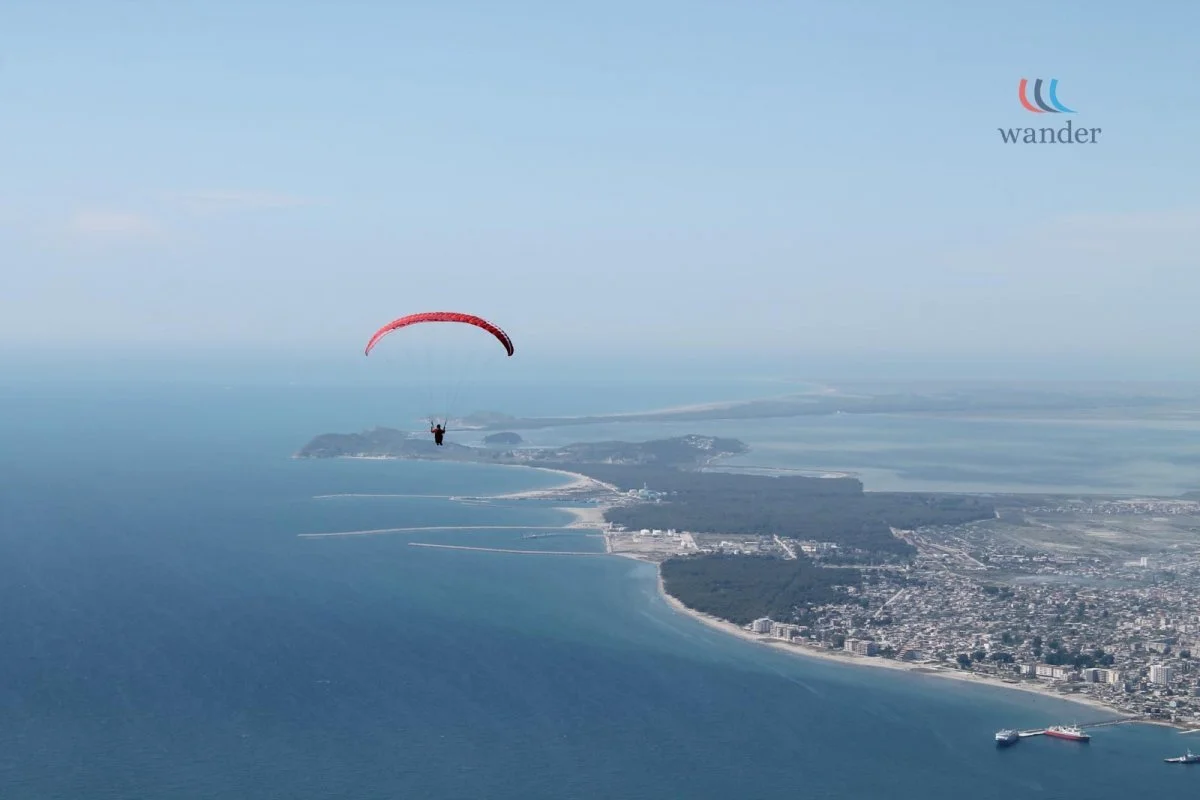 A person paragliding over a coastal city with buildings, shoreline, and ocean, with a cloudy blue sky in the background.