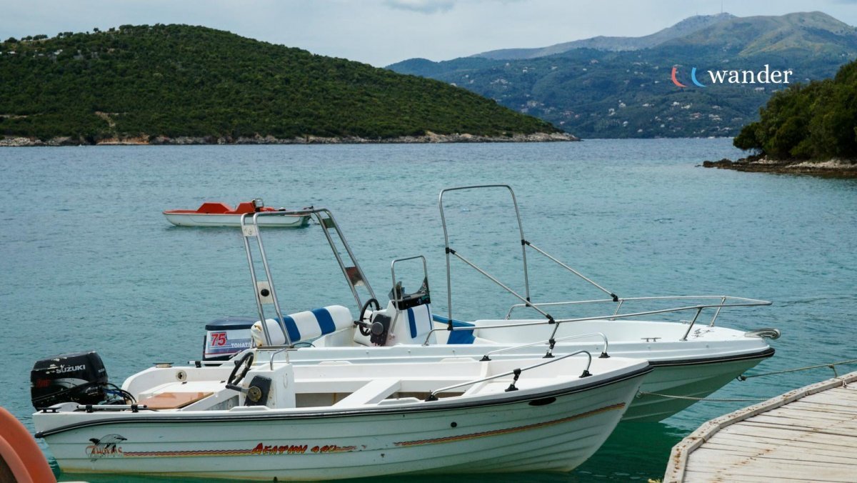 Two boats docked by a wooden pier on a calm body of water with green hills and mountains in the background, and a small red and white boat floating further out.