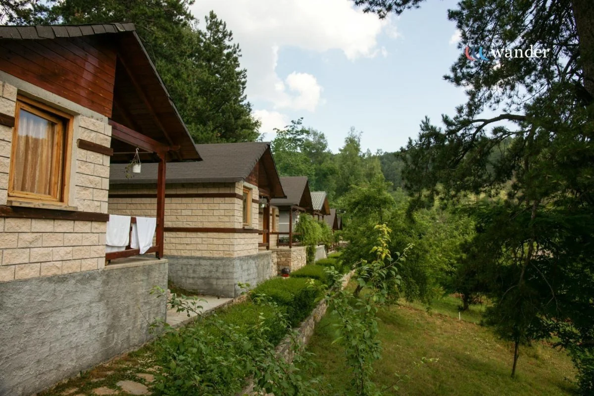 A row of small houses with brick and wood exteriors, each with a small porch and a window. Green trees and grass surround the houses, and the sky is partly cloudy.