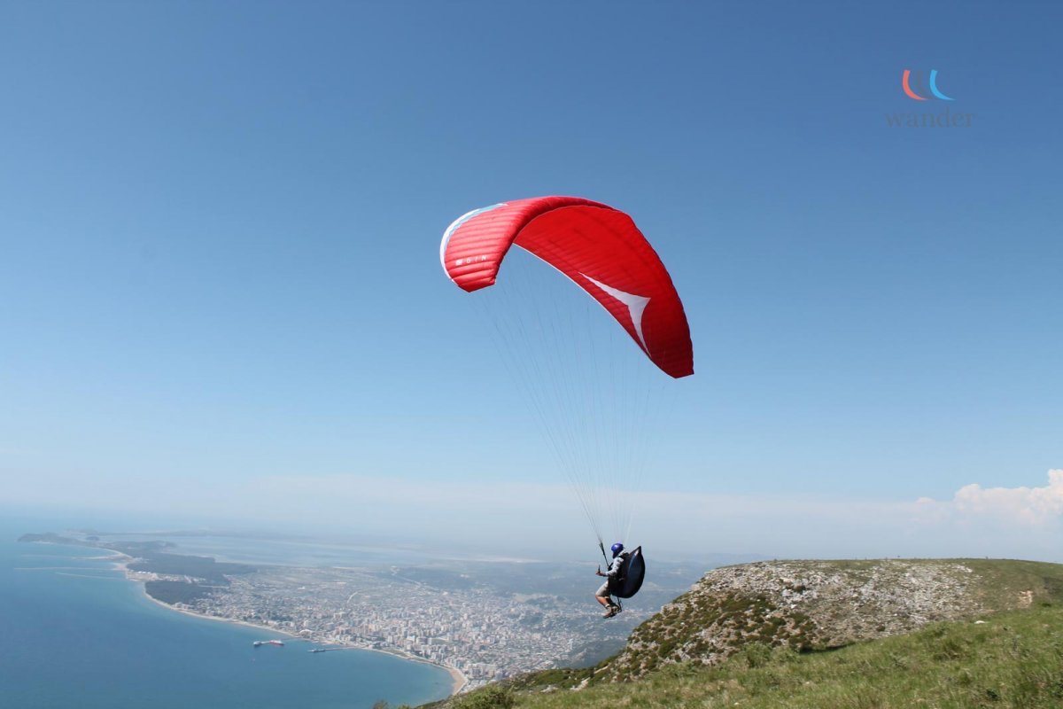 Person paragliding over a coastline with a city in the distance, under a clear blue sky.