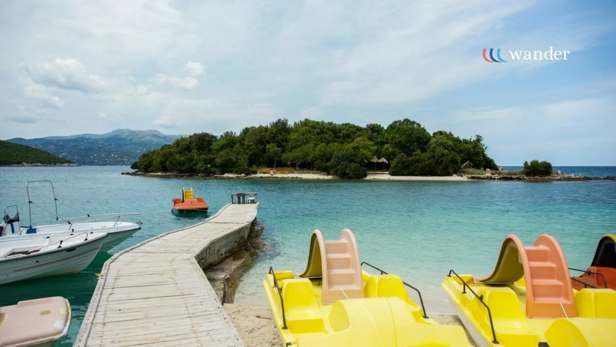 Tropical beach scene with a wooden dock leading to the water, paddle boats, and a lush island in the background under a partly cloudy sky.