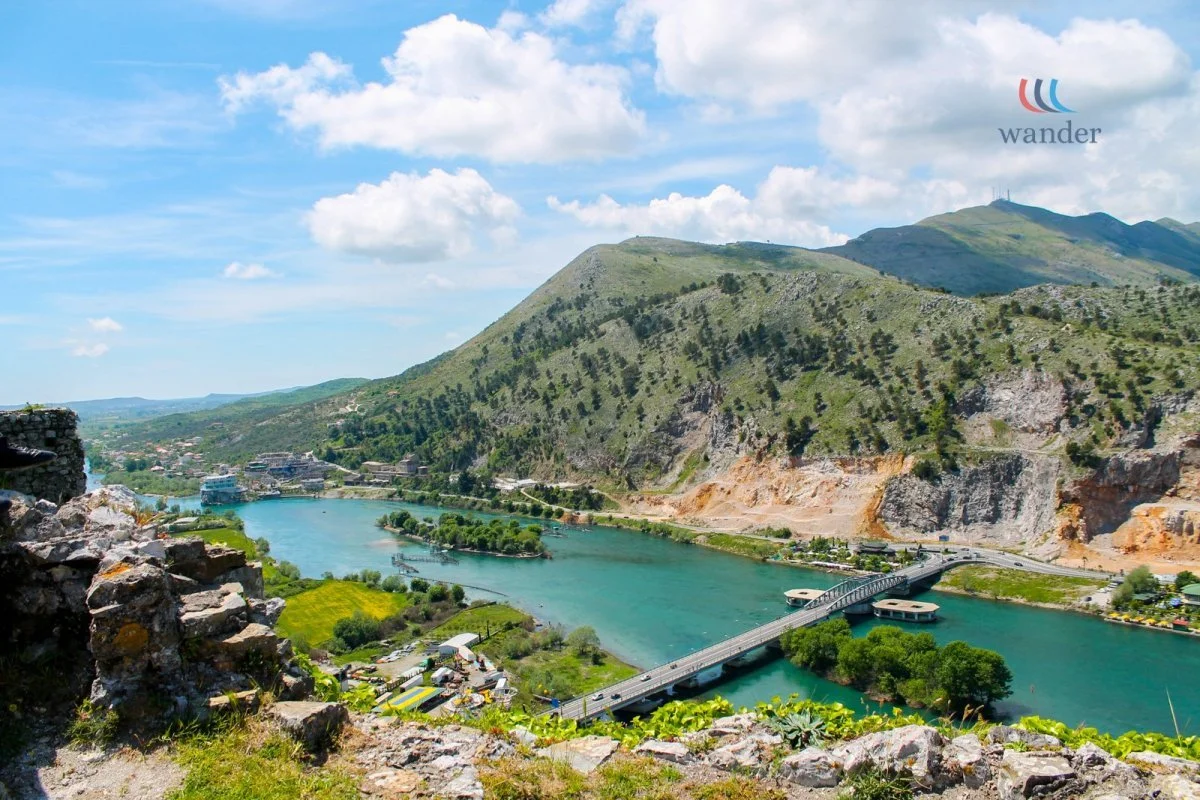 Scenic view of a river flowing through a valley with green hills and mountains in the background, a bridge crossing the river, and a small town on the riverbank.