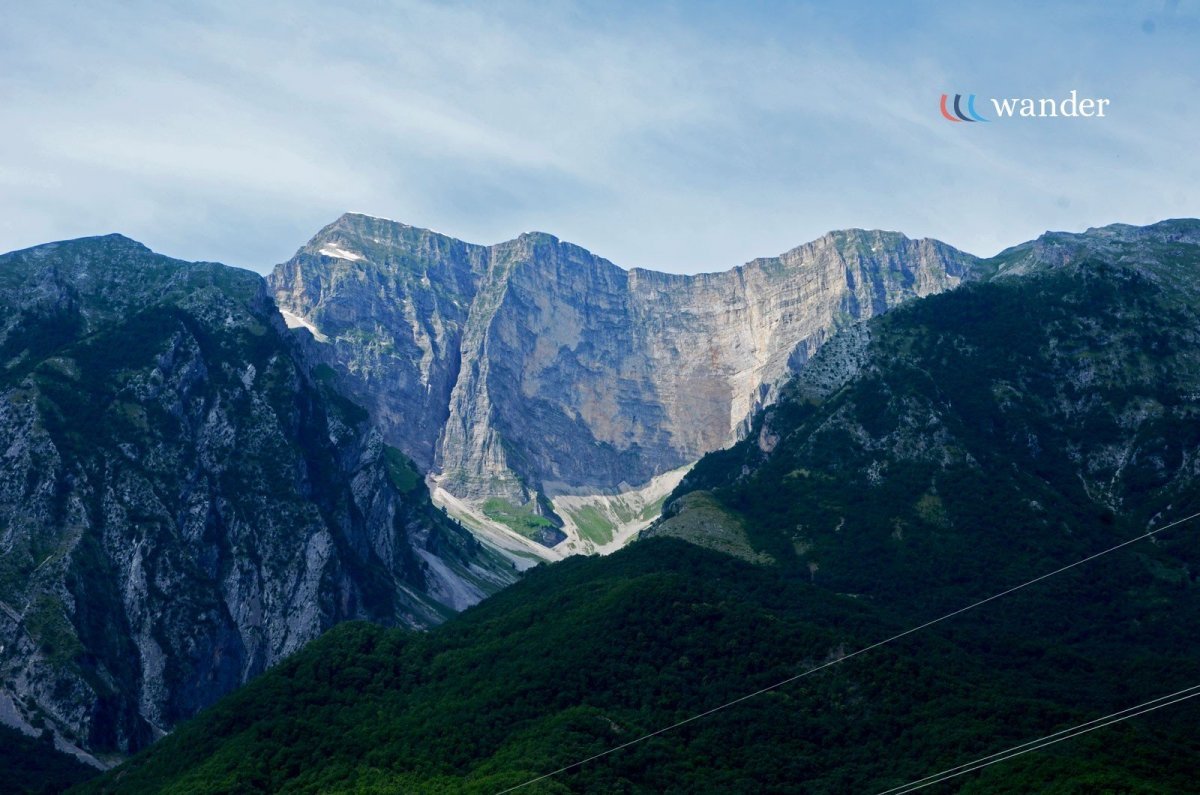 A mountain landscape with rocky peaks and green forested slopes under a cloudy blue sky.