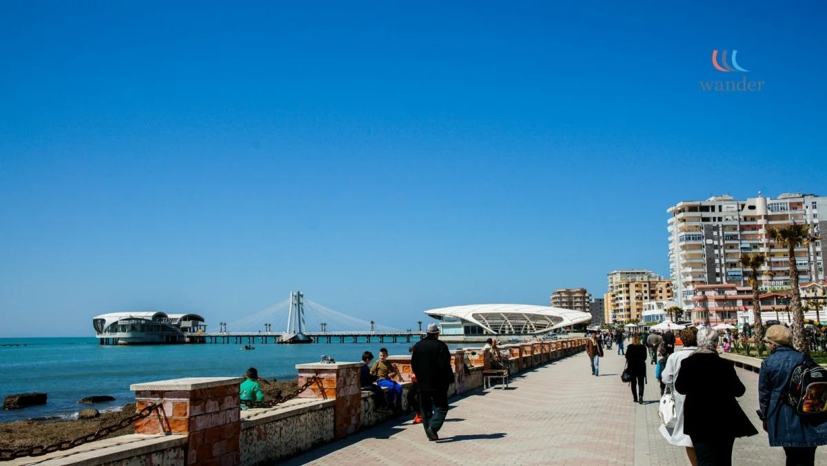People walking along a seaside promenade with modern buildings and a bridge in the background.