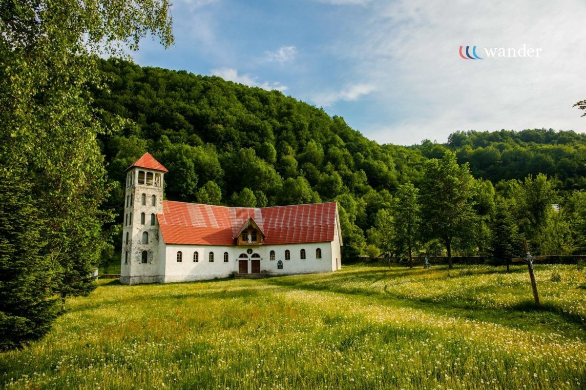 A white church with a red roof and a bell tower, situated on a grassy field surrounded by trees and a forested hillside, under a partly cloudy sky.