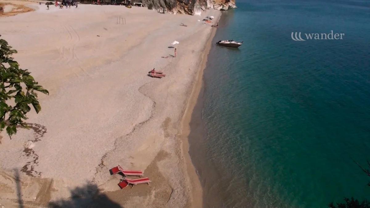 Beach with lounge chairs and umbrellas along the shore, a boat anchored in the water, and a cliff in the background.