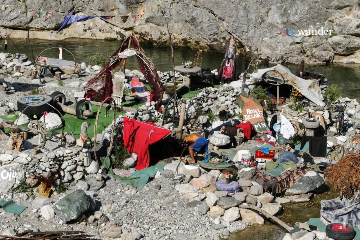 A makeshift outdoor campsite with various tents and shelters built from rocks, fabric, and other materials near a river. Items like tires, clothes, and supplies are scattered around, and a person is seen preparing or organizing belongings amidst the debris.