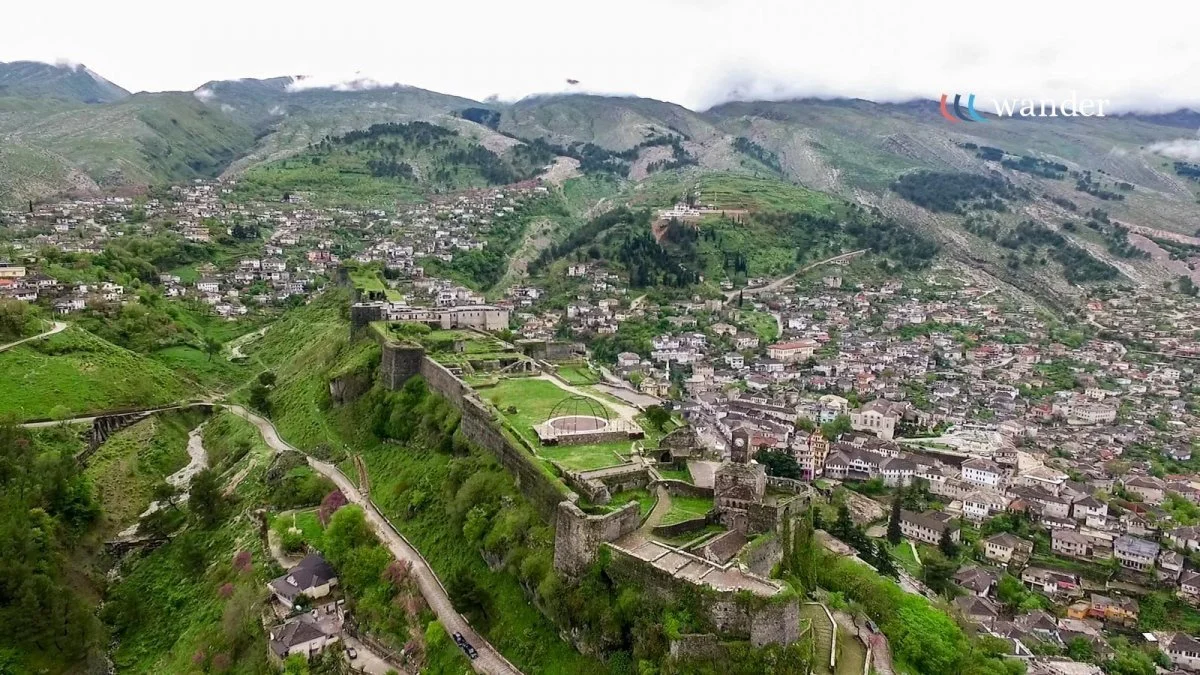 Aerial view of a hillside town with a historic castle and fortress ruins, surrounded by lush green mountains and buildings.