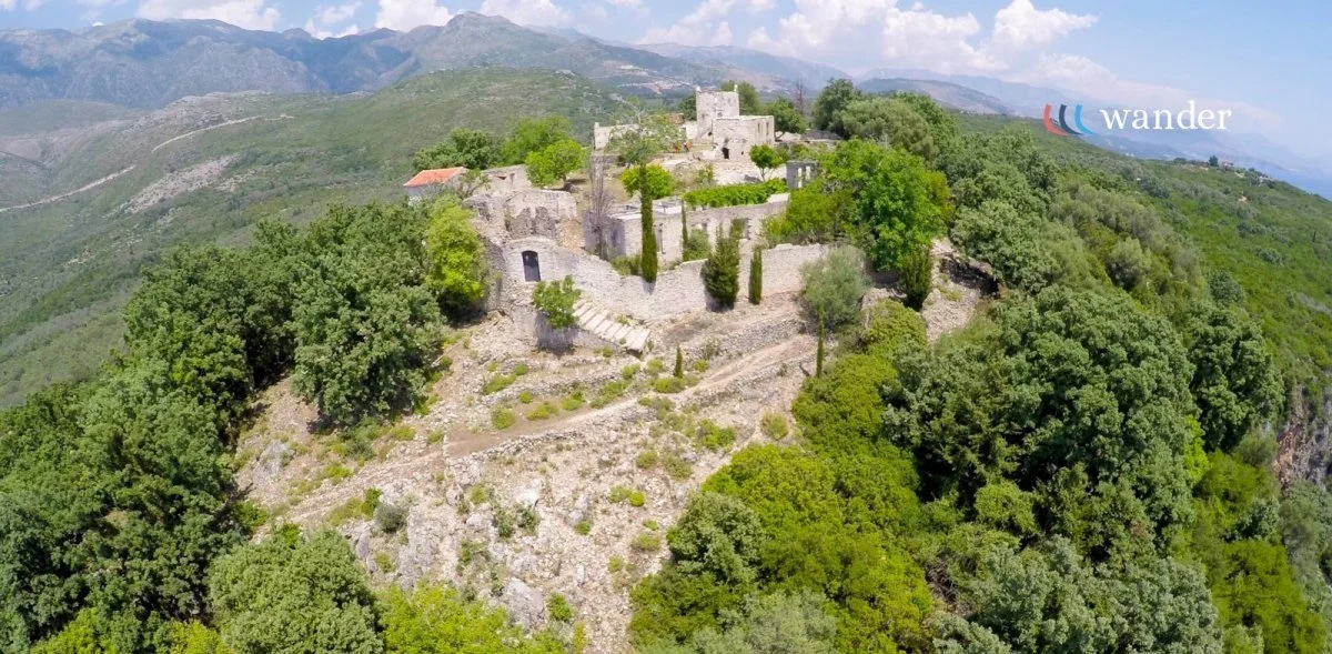 Aerial view of an ancient stone castle surrounded by lush green trees on a hillside with mountains in the background.