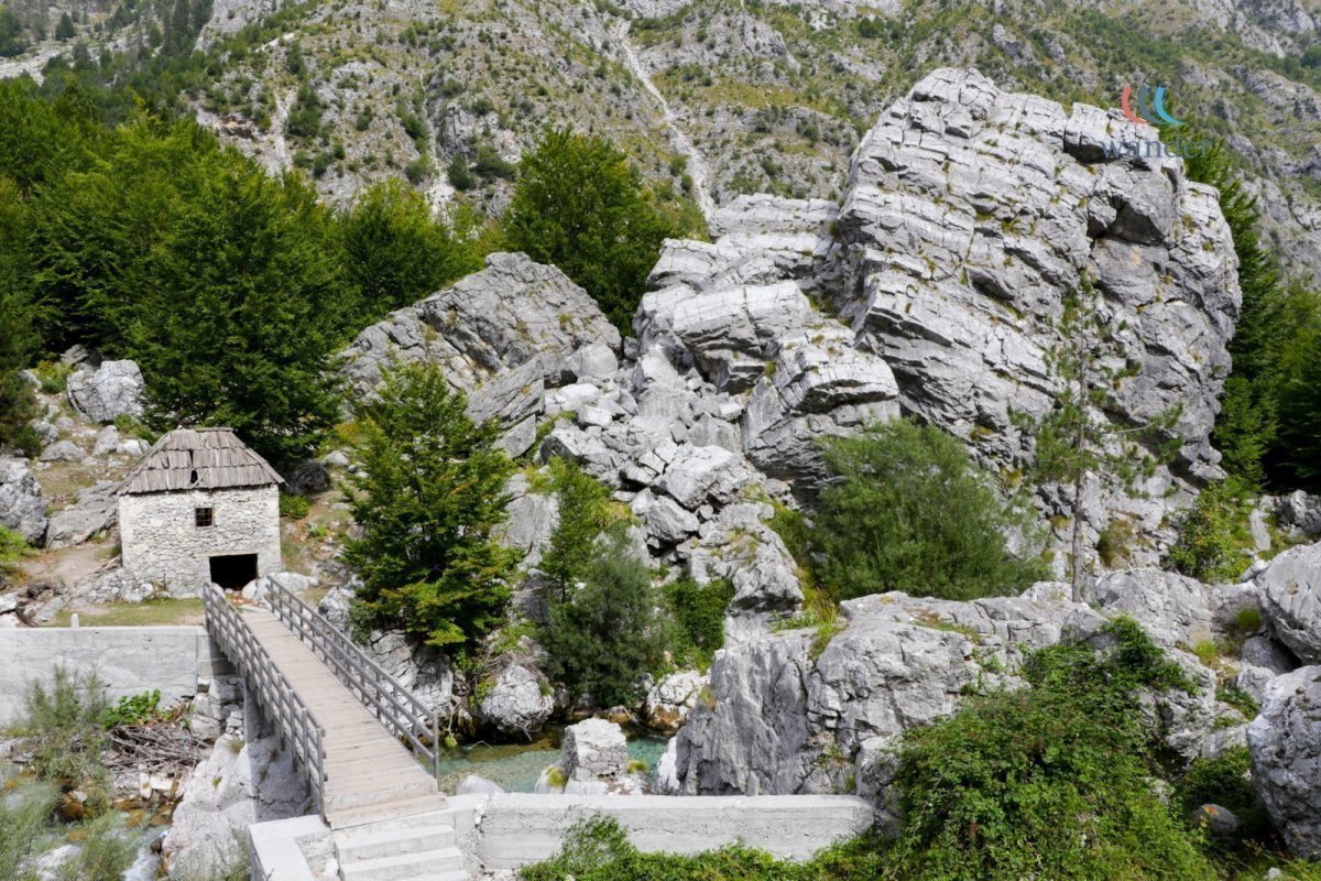 A small stone house with a wooden roof located near a wooden bridge, surrounded by large rocks and green trees in a mountainous area.