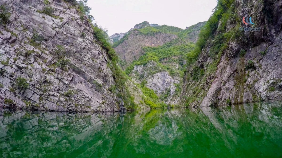 A narrow canyon with steep rocky walls covered in greenery, filled with calm green water reflecting the landscape.