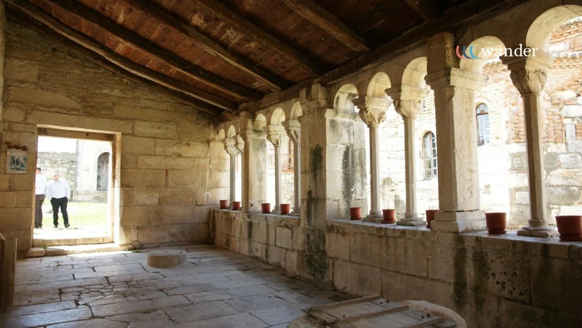 Interior of an ancient stone building with a wooden ceiling and a row of stone columns with arches, overlooking a courtyard. Red flower pots are placed along the window ledge.