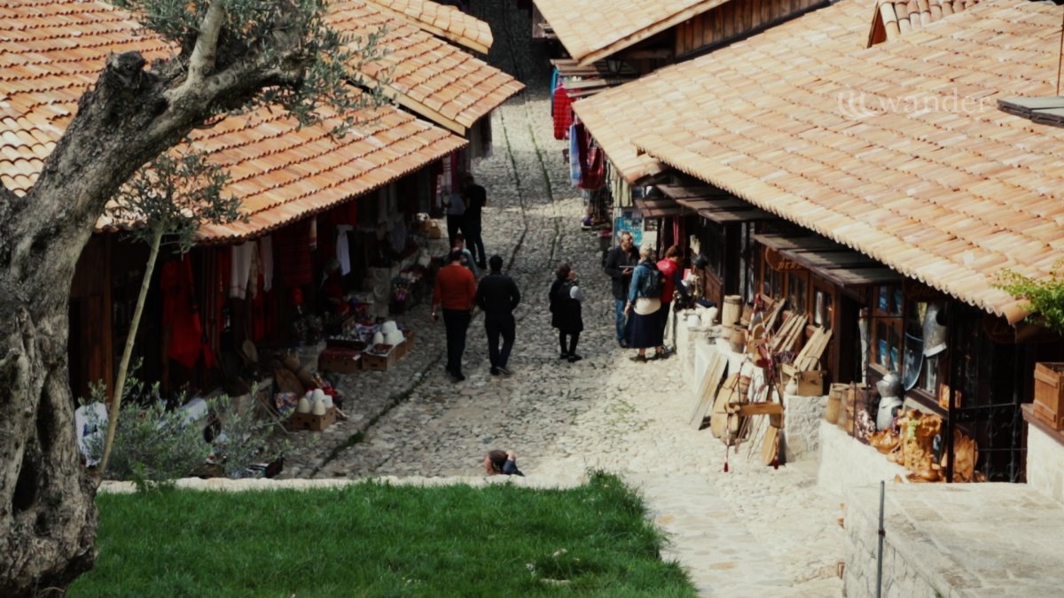 A cobblestone street market with vendors and shoppers, lined with small shops and orange-tiled roofs, in a historic area.
