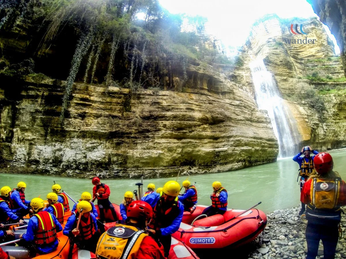 Group of people in helmets and life jackets preparing for a river adventure near a waterfall in a canyon.