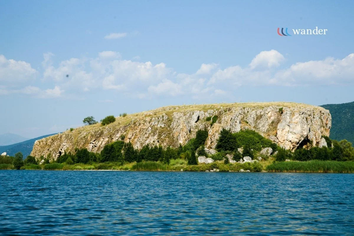 A large rocky hill or small mountain with green vegetation at its base, situated by a body of water with a blue sky and white clouds overhead.