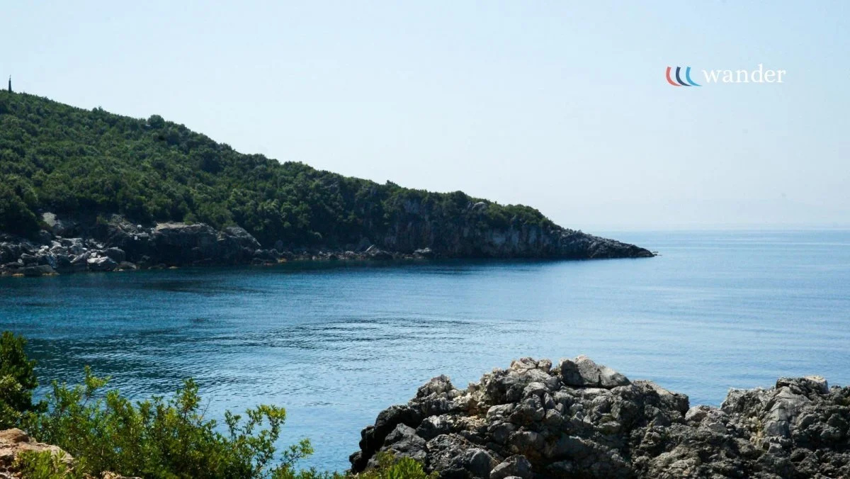 A serene coastal scene with clear blue water, rocky shoreline in the foreground, and green forested hills in the background under a pale blue sky.