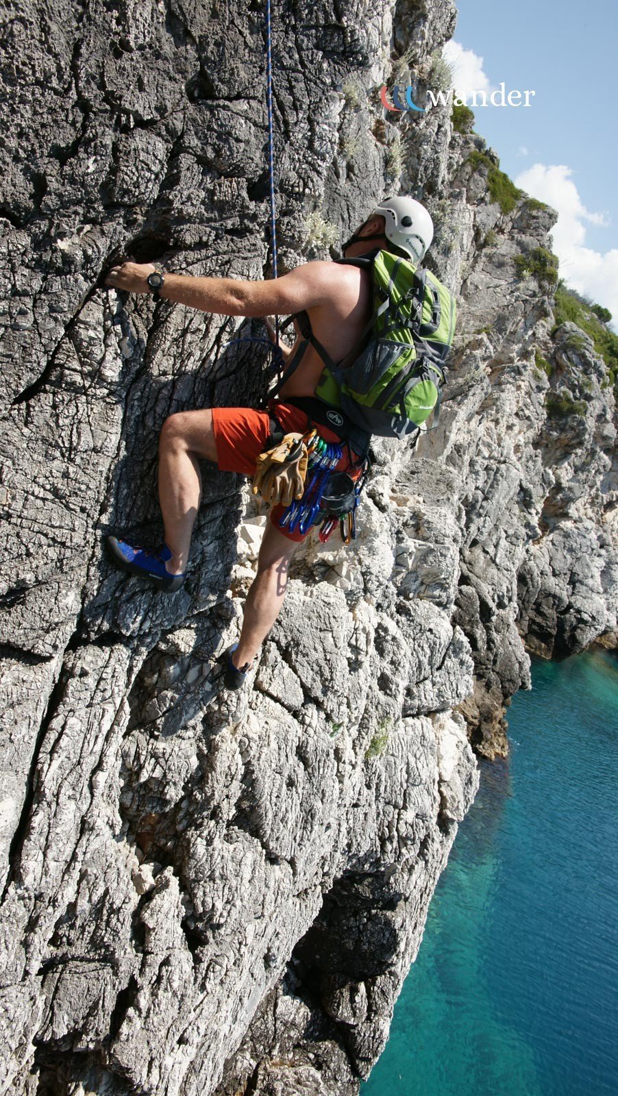 A male rock climber wearing a helmet and red shorts climbing a rugged limestone cliff near water, with climbing gear attached to his waist and a backpack, during daytime.