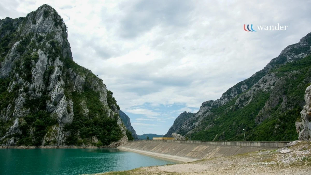 A reservoir between green, rocky mountains with a concrete dam and cloudy sky.