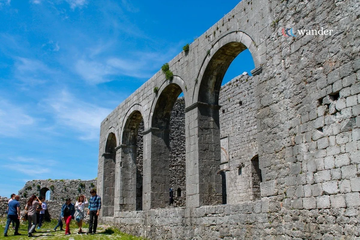 Ancient stone ruins with large arches and a group of people walking nearby under a bright blue sky.