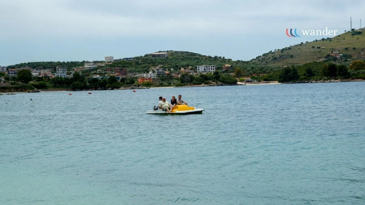 Four people riding a yellow paddle boat on a lake with a town and green hills in the background, under a cloudy sky.