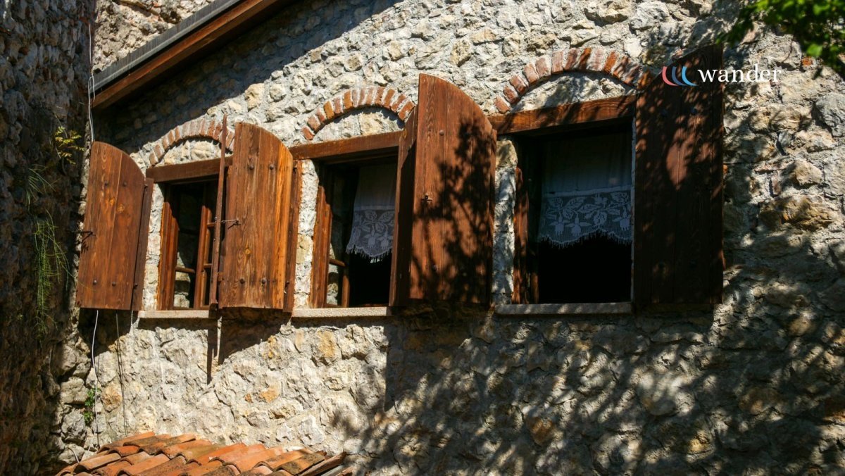 Stone house with three small windows and wooden shutters, partially open, with lace curtains inside. Shadows of tree branches cast on the wall.