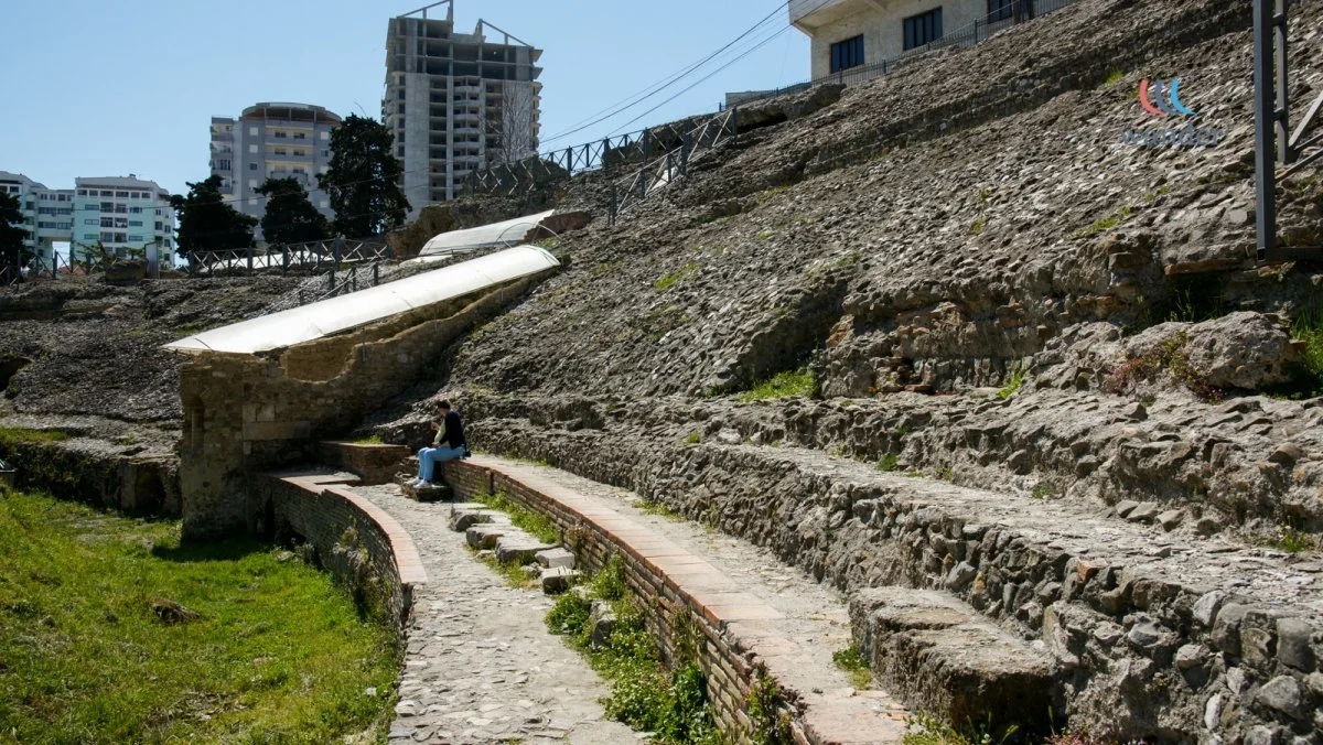 Person sitting on a stone bench on an ancient archaeological site with stone steps and ruins, with modern buildings in the background under a clear blue sky.