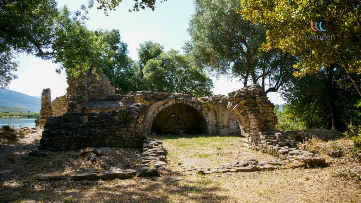 Ruins of an ancient stone structure with arches, surrounded by trees and near a body of water.