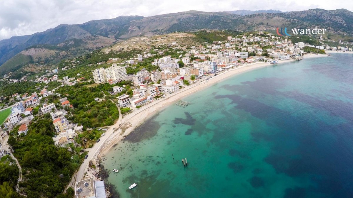 Aerial view of a coastal town with a sandy beach and turquoise water, mountains in the background, and buildings along the shoreline. The image has a logo with the word "wander" and colorful curved lines.