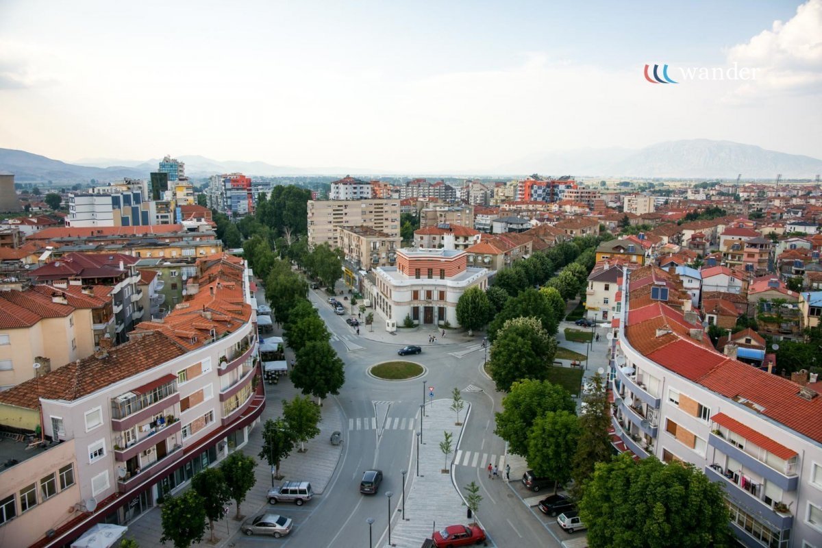 Cityscape with residential buildings, trees, streets, and distant mountains under a partly cloudy sky.
