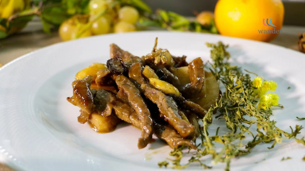 A white plate with a serving of cooked eggplant with a glossy sauce, garnished with herbs and yellow flowers, with grapes and an orange in the background.