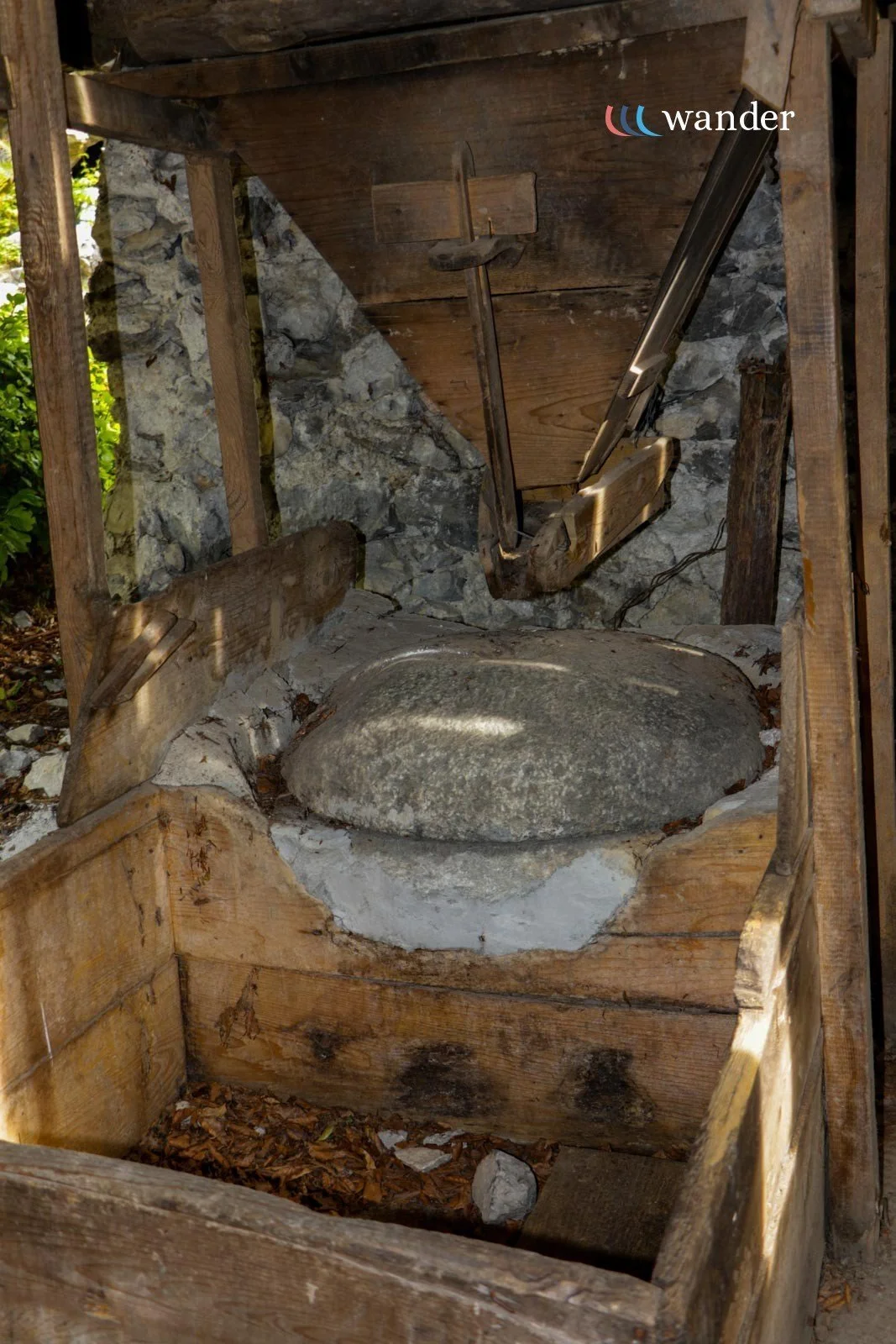 A primitive outdoor stone mill with a large rounded stone wheel inside a wooden frame, used for grinding or crushing, situated outdoors with stone and wood construction.