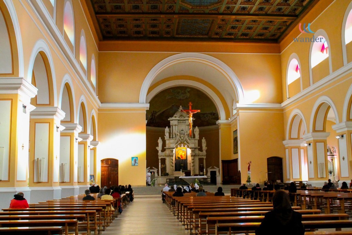 Interior of a church with rows of wooden pews, a high ceiling with ornate decorations, and an altar with a crucifix and religious icons at the front.
