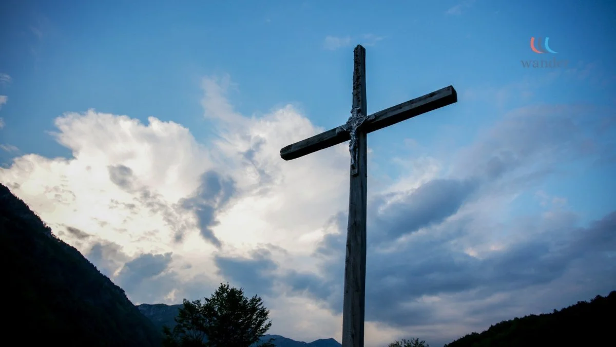 A wooden cross stands outdoors against a cloudy sky and mountain landscape.