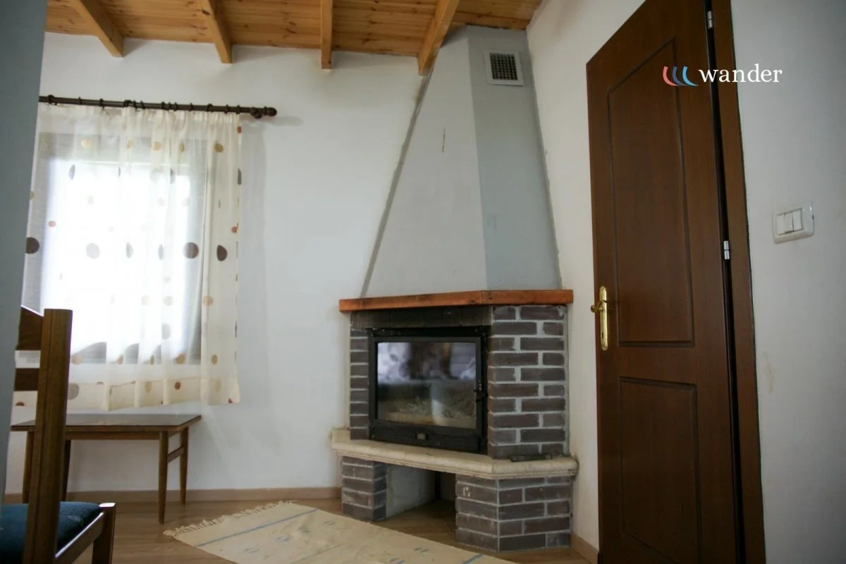 Living room with a window, curtain with colorful polka dots, a brick fireplace with a glass door, wooden mantel, and a closed wooden door. The ceiling has exposed wooden beams.