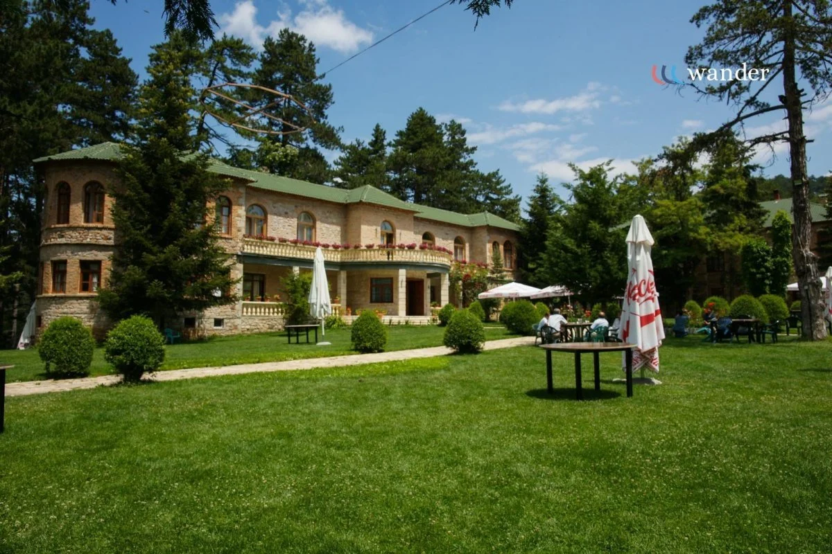 A large stone house with a green roof surrounded by a well-maintained lawn, trees, and outdoor seating with umbrellas.