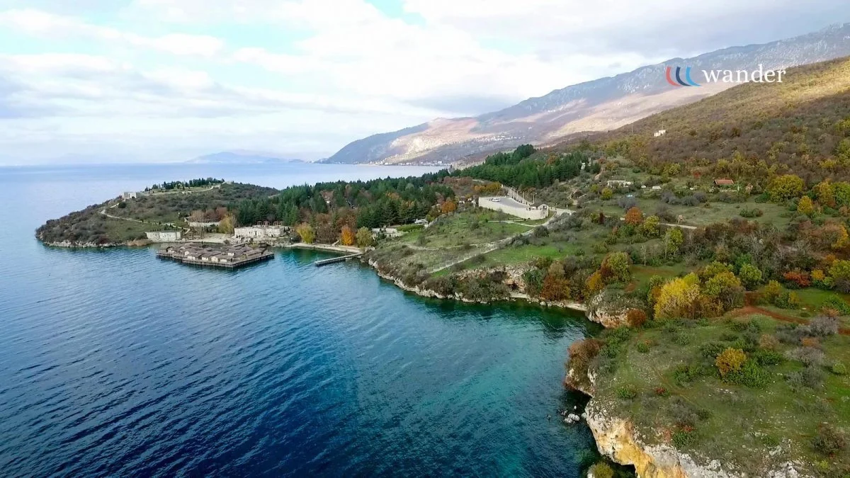 Aerial view of a coastal landscape with a shoreline, trees, houses on a hillside, and mountains in the background.