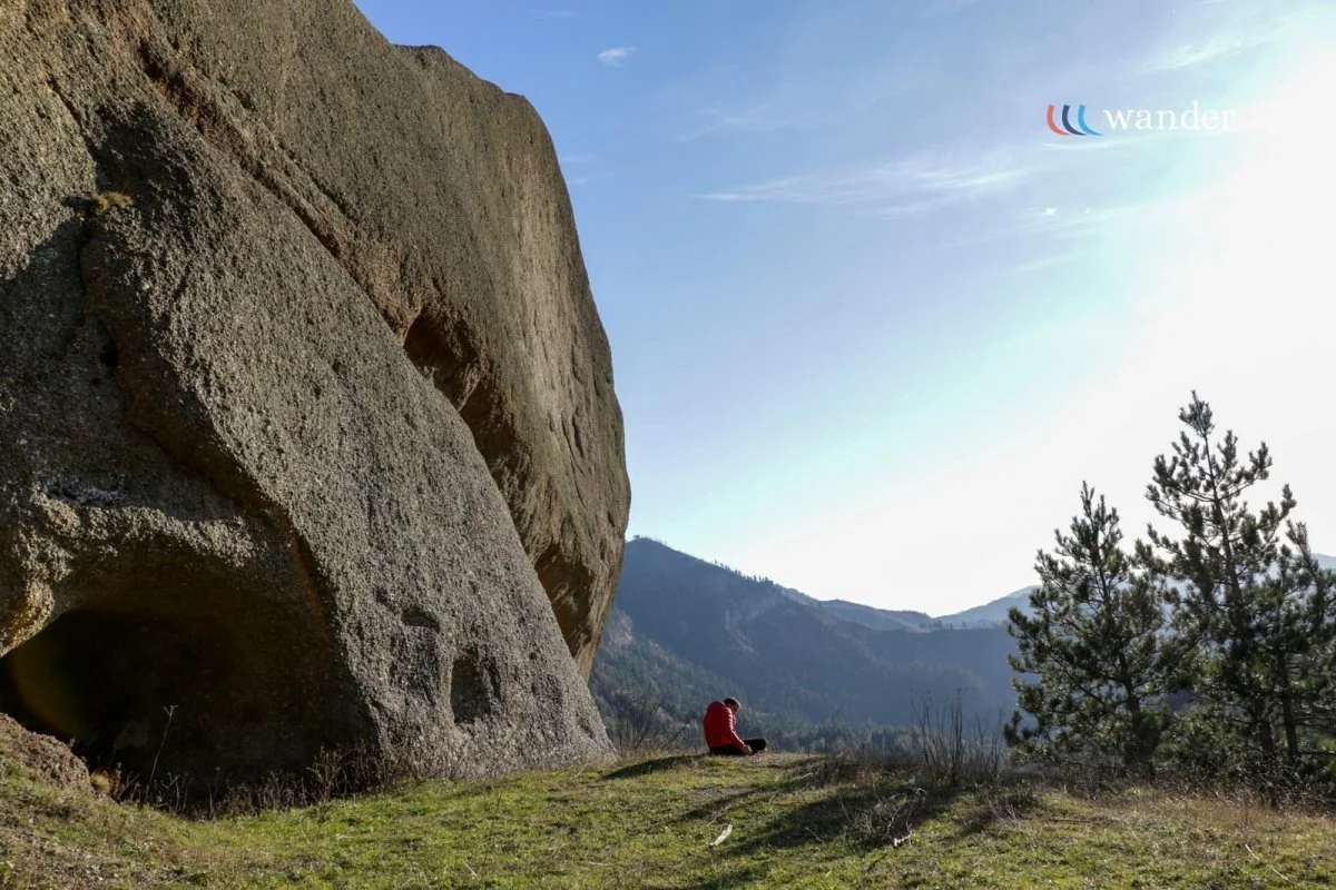 A person in a red jacket sitting on the grass near a large rock formation in a mountainous area, with trees and hills in the background under a clear blue sky.
