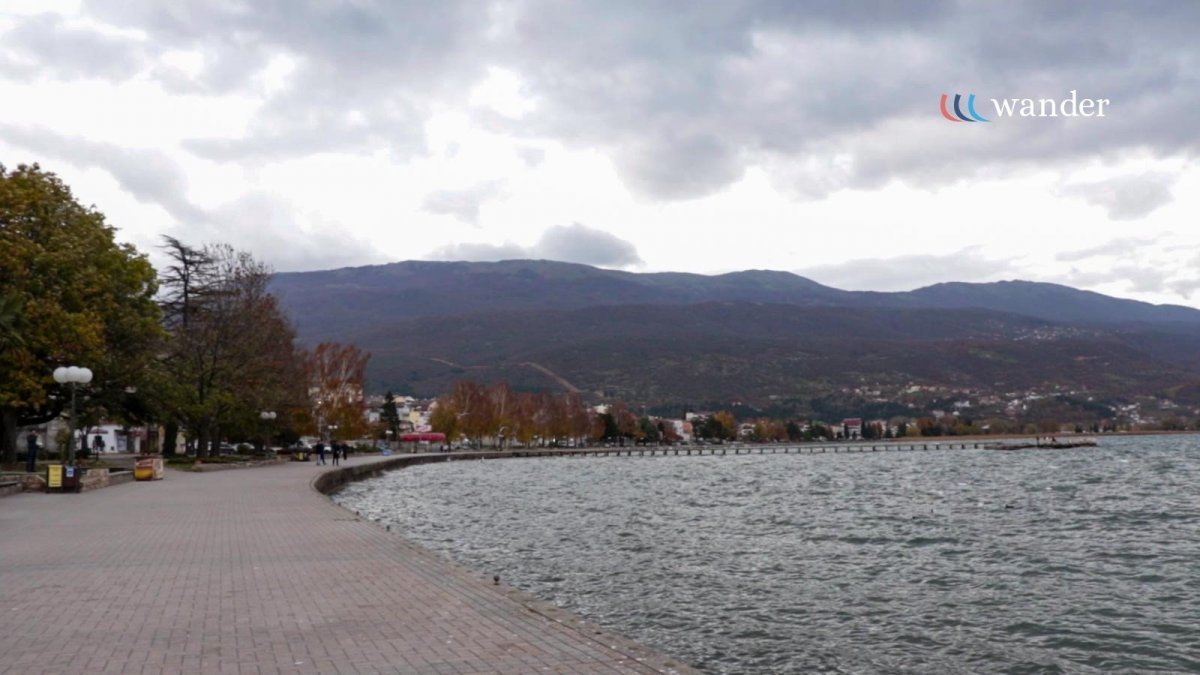 A lakeside promenade with a paved walkway, trees, and lampposts, overlooking a body of water with mountains in the background, under a cloudy sky.