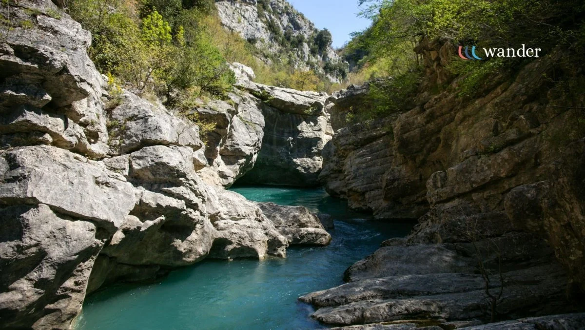 A narrow canyon with large gray rocks and green trees on either side, featuring a small waterfall and a turquoise river flowing through it.