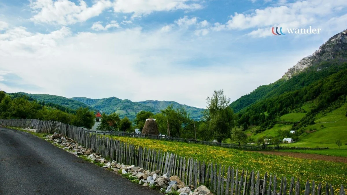Scenic rural landscape with green hills, mountains, a blue sky with clouds, a dirt path with rocky edges, a wooden fence, farm buildings, and trees.