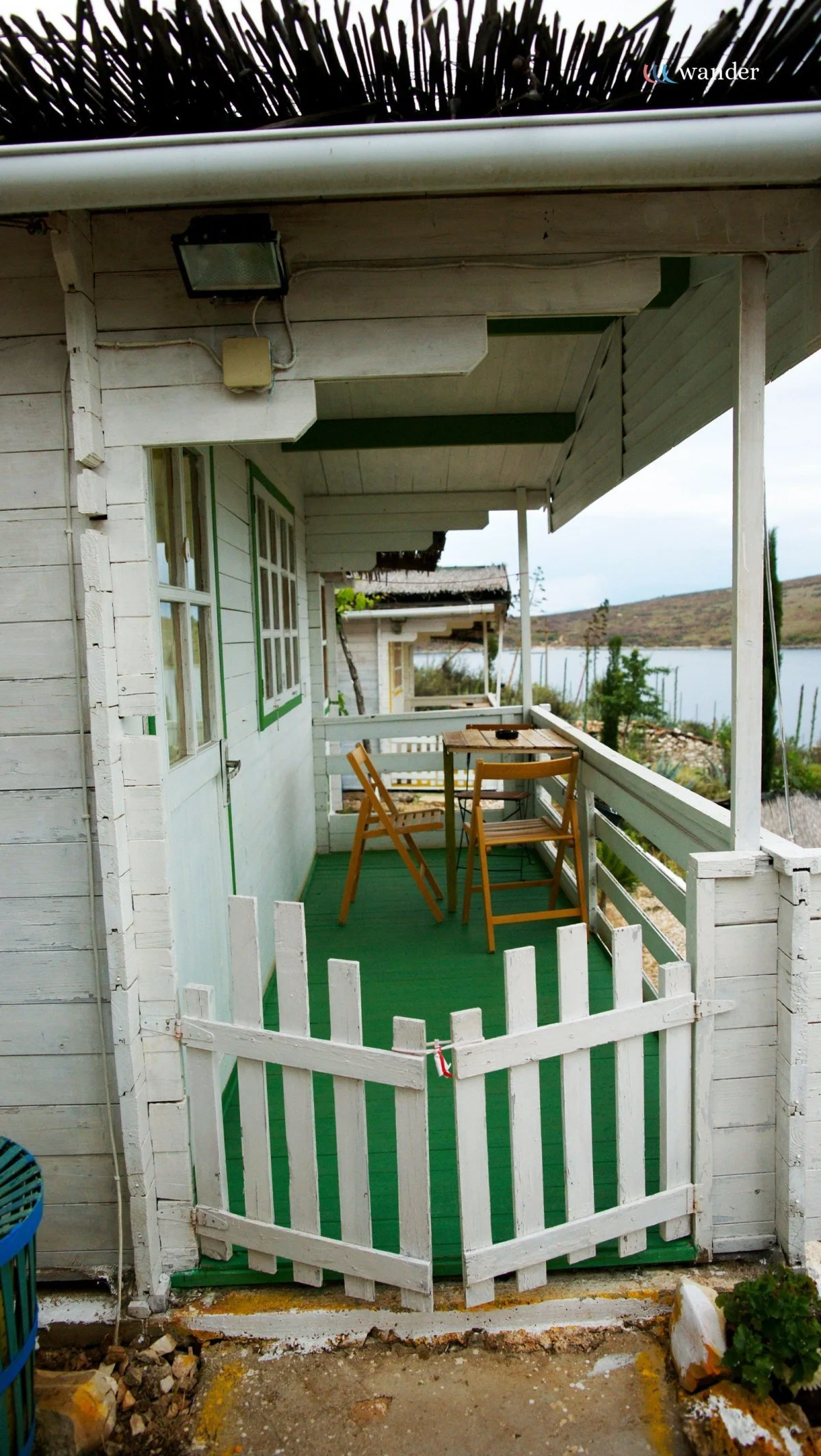 A small, rustic white painted cottage with an outdoor balcony overlooking a body of water. The balcony has a small wooden table and two chairs, surrounded by a white wooden fence, with a view of hills and trees in the background.